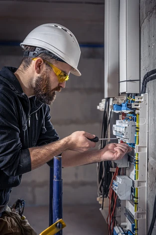 Électricien professionnel travaillant sur un tableau électrique dans un bâtiment en construction. L'homme porte un casque de sécurité blanc, des lunettes de protection jaunes et manipule des câbles avec précision.