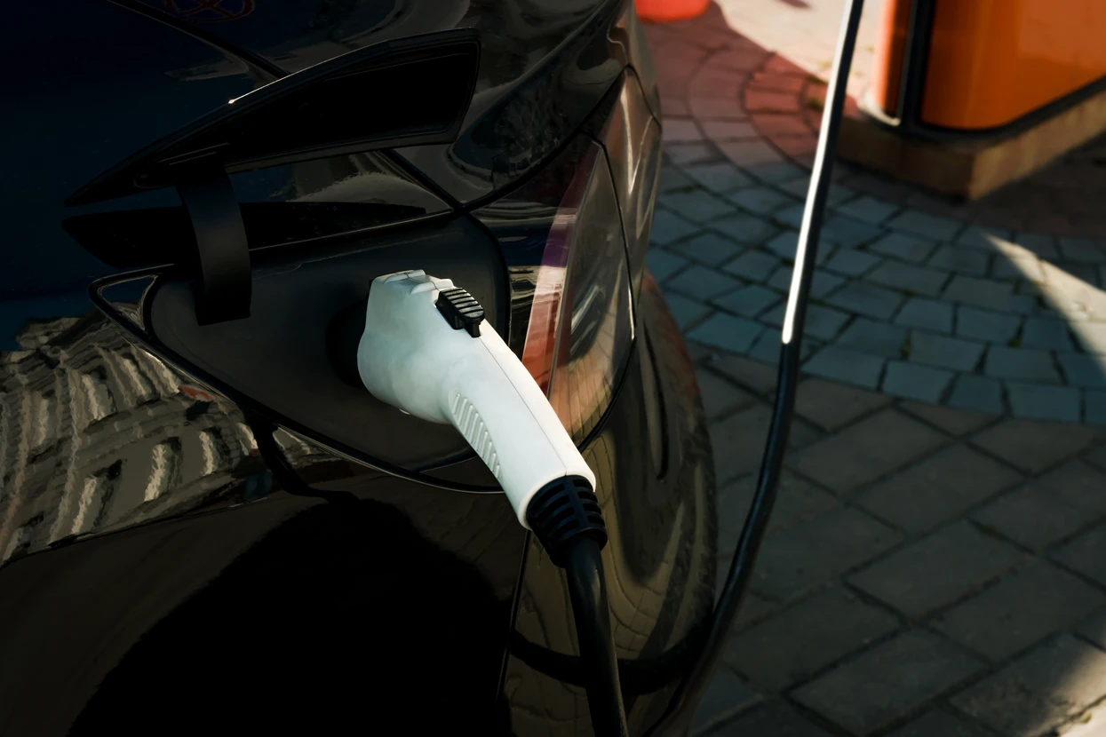 Scène extérieure urbaine avec véhicule électrique en charge sur pavés. Un conducteur connecte un câble de recharge blanc à la prise du véhicule sombre.