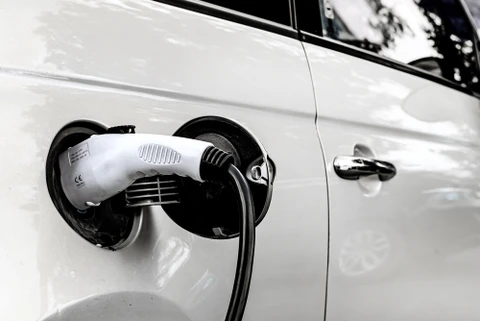 Extérieur jour, véhicule blanc en charge sur station électrique. Câble d'alimentation connecté au port de recharge latéral, portière conducteur visible.