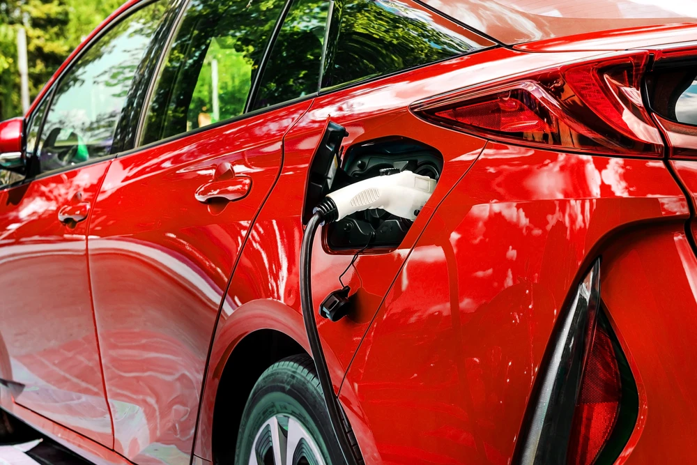 Véhicule électrique rouge en charge à l'extérieur, connecteur blanc inséré dans la prise de recharge latérale, environnement verdoyant visible en arrière-plan.