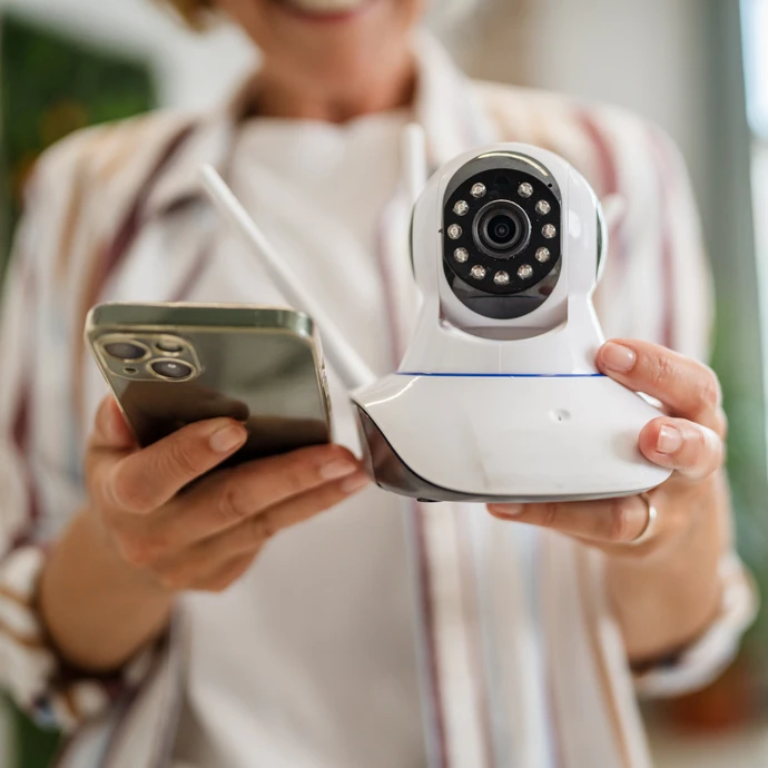 Un technicien en uniforme rouge installe une cam&eacute;ra de surveillance au plafond d'un int&eacute;rieur aux murs blancs. L'homme porte une casquette et des gants de protection rouges, utilisant un tournevis pour fixer le dispositif de s&eacute;curit&eacute;.
